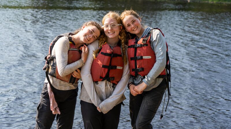 Three young women are standing close together, smiling at the camera, with a body of water in the background. They are all wearing life jackets, suggesting they may be about to engage in water activities. The woman in the center wears glasses. The water behind them is slightly choppy, indicating a possible breeze.